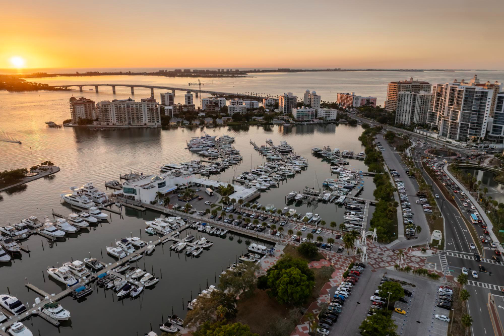 Aerial marina with boats, waterfront buildings, bridge at sunset.